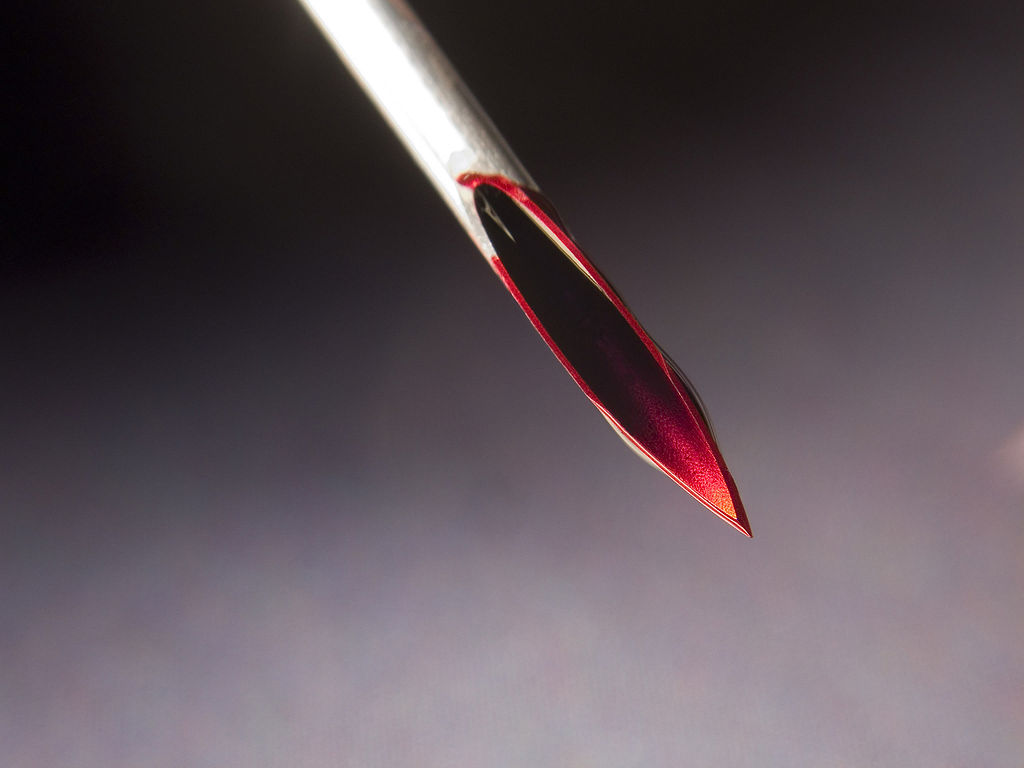 toronto play piercing workshop needle play 101 - Image description: a close up shot of a hollow needle with blood all over the tip, set against a background that fades from black to grey