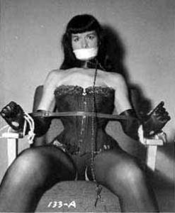 bdsm basics peterborough kink classes kinky workshops youre welcome ptbo [Image Description: Betty page, a white woman with long black hair and bangs sits bound to a chair by rope and a spreader bar. she has tape over her mouth and is dressed in leather lingerie]