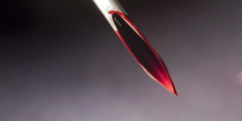 toronto play piercing workshop needle play 101 - Image description: a close up shot of a hollow needle with blood all over the tip, set against a background that fades from black to grey